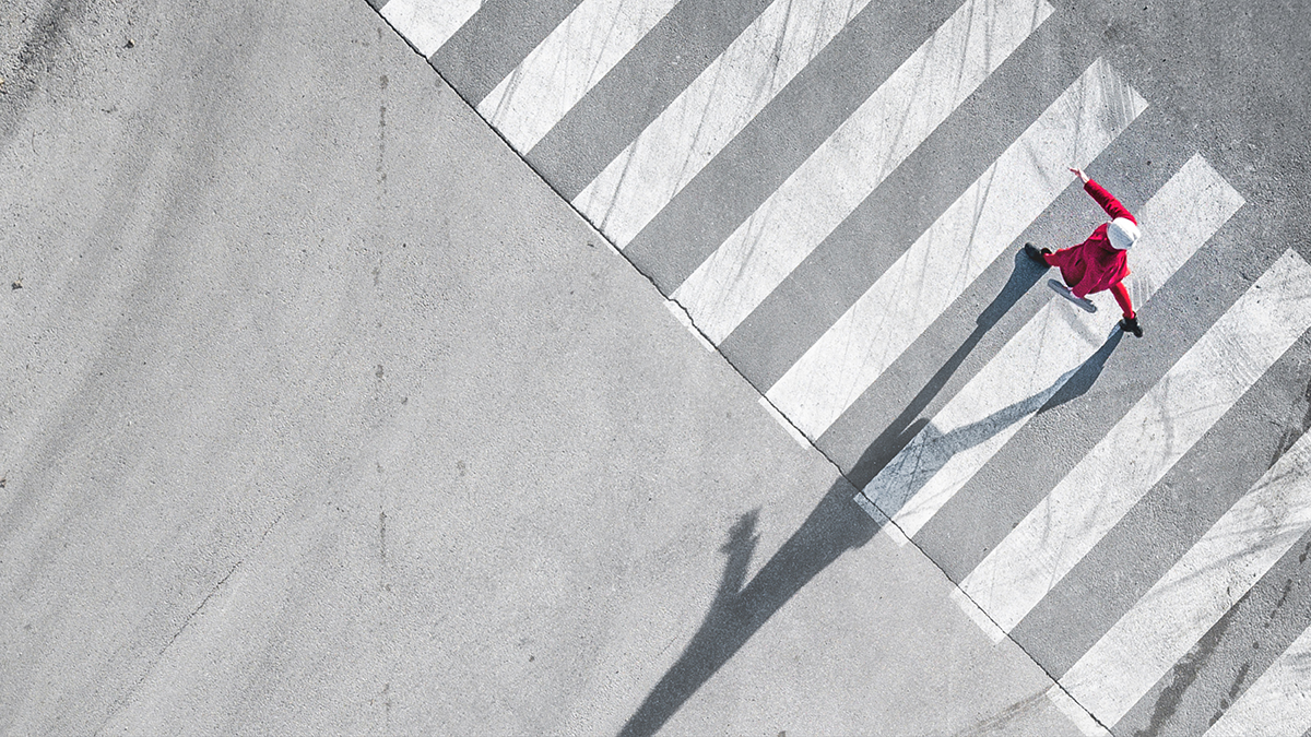 Eine einzelne Person von oben fotografiert. Sie geht auf der menschenleeren Strasse einer internationalen Grossstadt &uuml;ber den Fussg&auml;ngerstreifen. Ihr Schatten wird von der Sonne als &uuml;bergrosse Silhouette auf die Strasse projiziert.