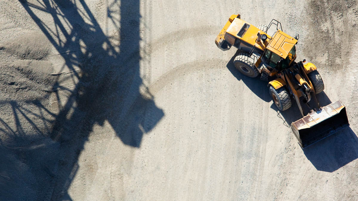 Blick von oben auf einen schweren Bagger, der im Kies-Sand-Boden scharf rechts wendet. Seine Spuren kreuzen sich mit den dunklen Schatten einer grossindustriellen Anlage.