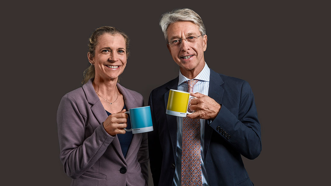 Verwaltungsratspräsident Herbert J. Scheidt und Verwaltungsrätin Maja Baumann lachend mit farbigen Tassen in der Hand vor dunklem Hintergrund
