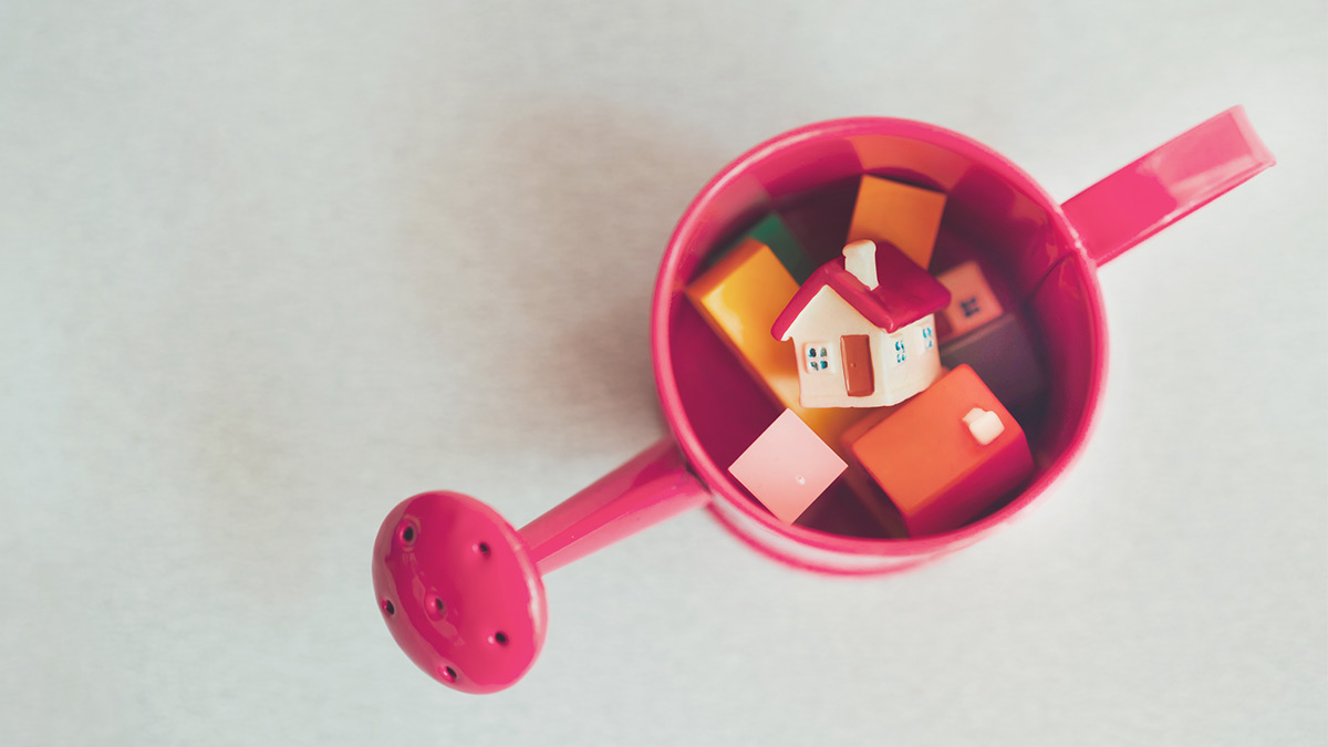 An analogy to the trickle-down-effect: Does with the UBI come “homeownership for all”? The picture shows a children’s watering can in red from above. Toy building blocks lie in it; one of them is a house with a gable roof.